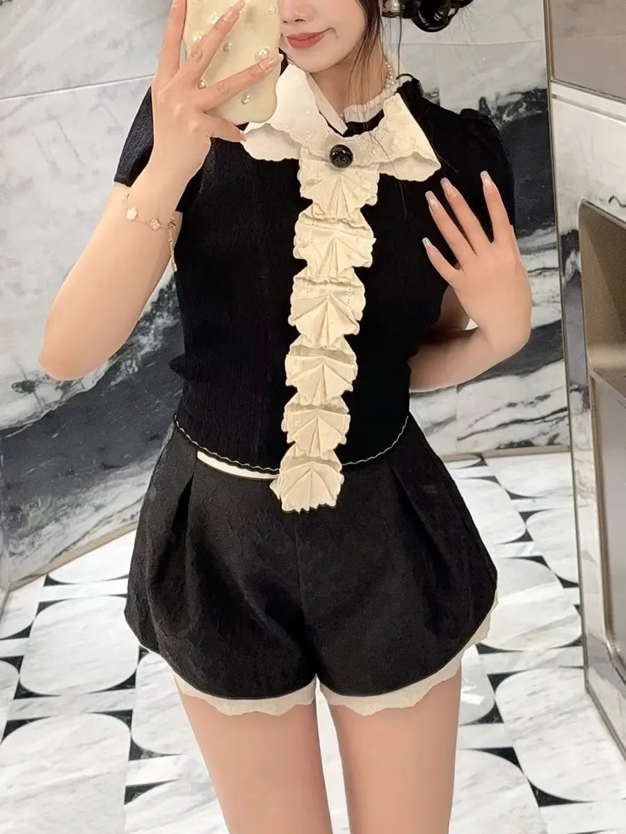 A woman models a chic black short-sleeve knit top with cascading white ruffles and a black bow detail, paired with matching black shorts featuring a white scalloped hem.