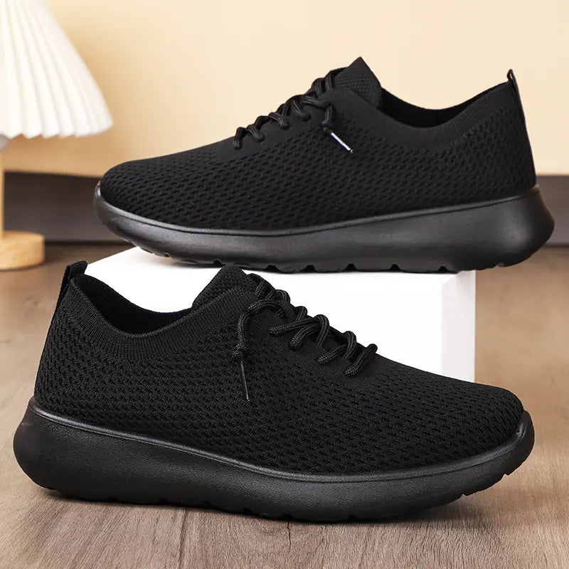 Two black engineered knit trainers shown from a three-quarter view, one elevated on a white cube, highlighting their breathable mesh uppers and lightweight EVA soles.