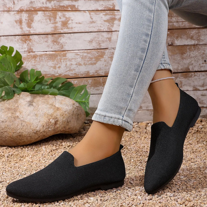 Black breathable knitted pointed toe flats worn by a woman in light wash jeans, styled with a pebble and greenery background.