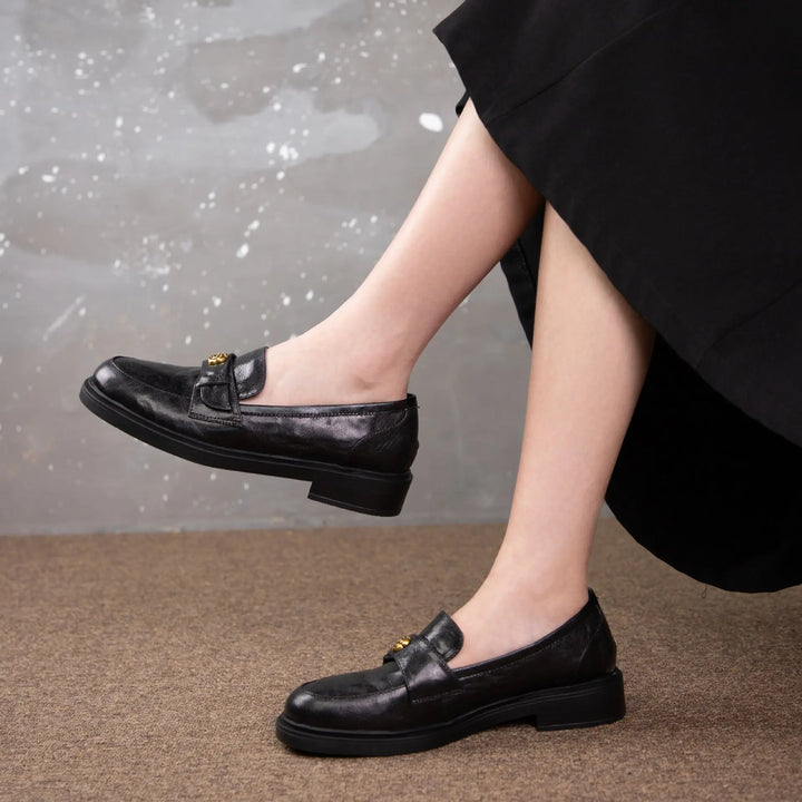 A pair of chic black leather loafers with a gold-toned floral buckle detail, shown on a woman&
