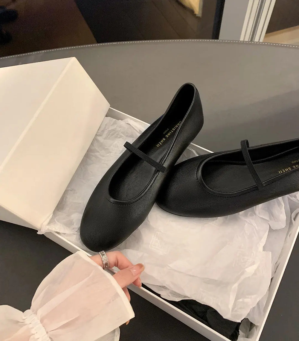 Black minimalist Mary Jane ballet flats with a thin elastic strap, presented inside a white box with crumpled tissue paper, alongside a hand with a ring.