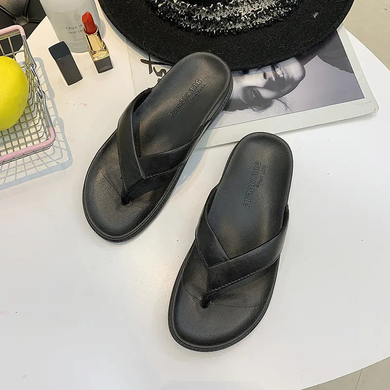 A pair of black matte faux leather flip-flop sandals with wide cross-straps and contoured footbeds, styled on a white table with accessories.