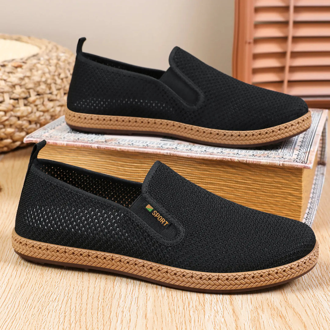 A pair of black men's breathable mesh slip-on loafers with a woven jute-like trim detail and a subtle 'SPORT' logo on the side, displayed on a wooden surface with a book and basket.