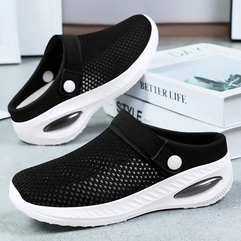 Black breathable mesh clogs with white air cushion soles and a decorative button strap, shown in a three-quarter side view. These slip-on shoes offer comfort and ventilation.