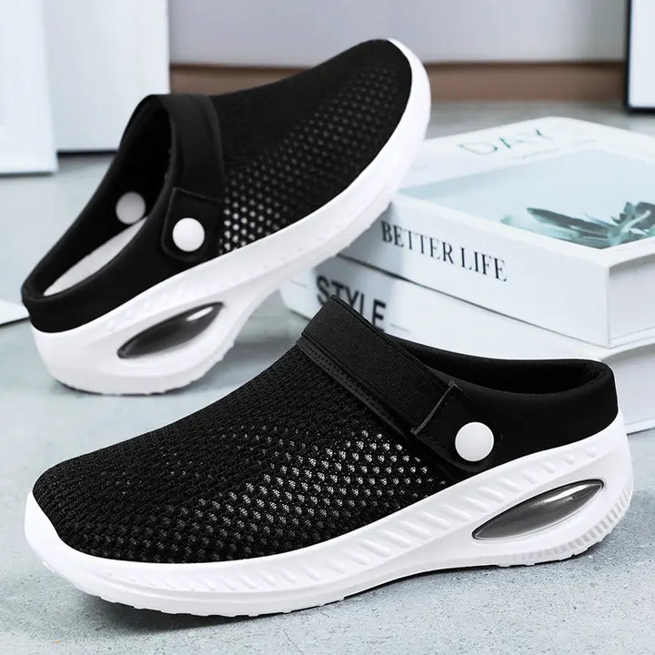 Black breathable mesh clogs with white air cushion soles and a decorative button strap, shown in a three-quarter side view. These slip-on shoes offer comfort and ventilation.
