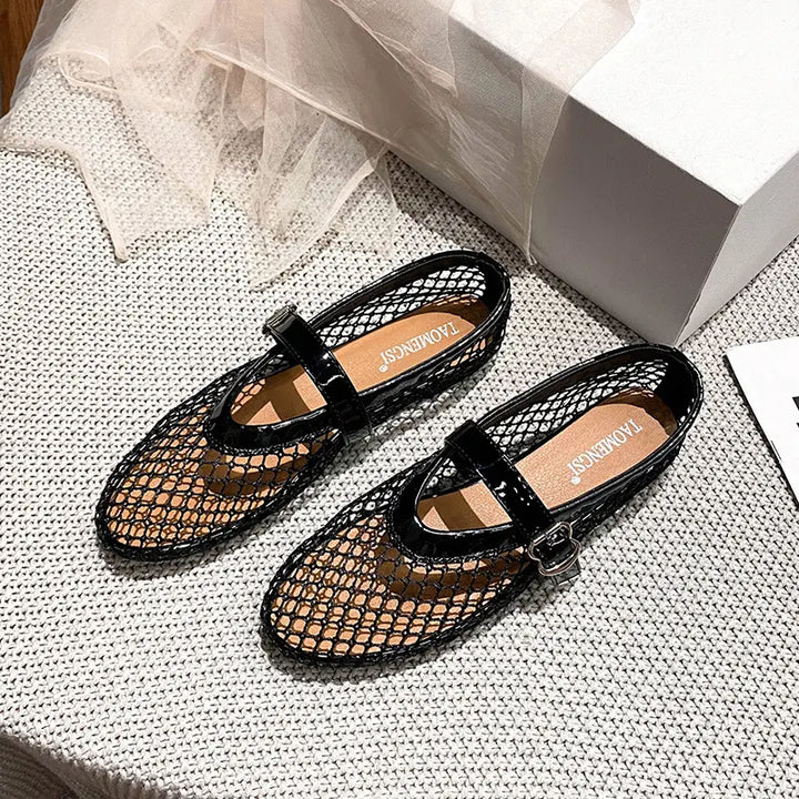 Black mesh ballet flats with patent Mary Jane strap and silver buckle, presented from a three-quarter angle on a textured surface.