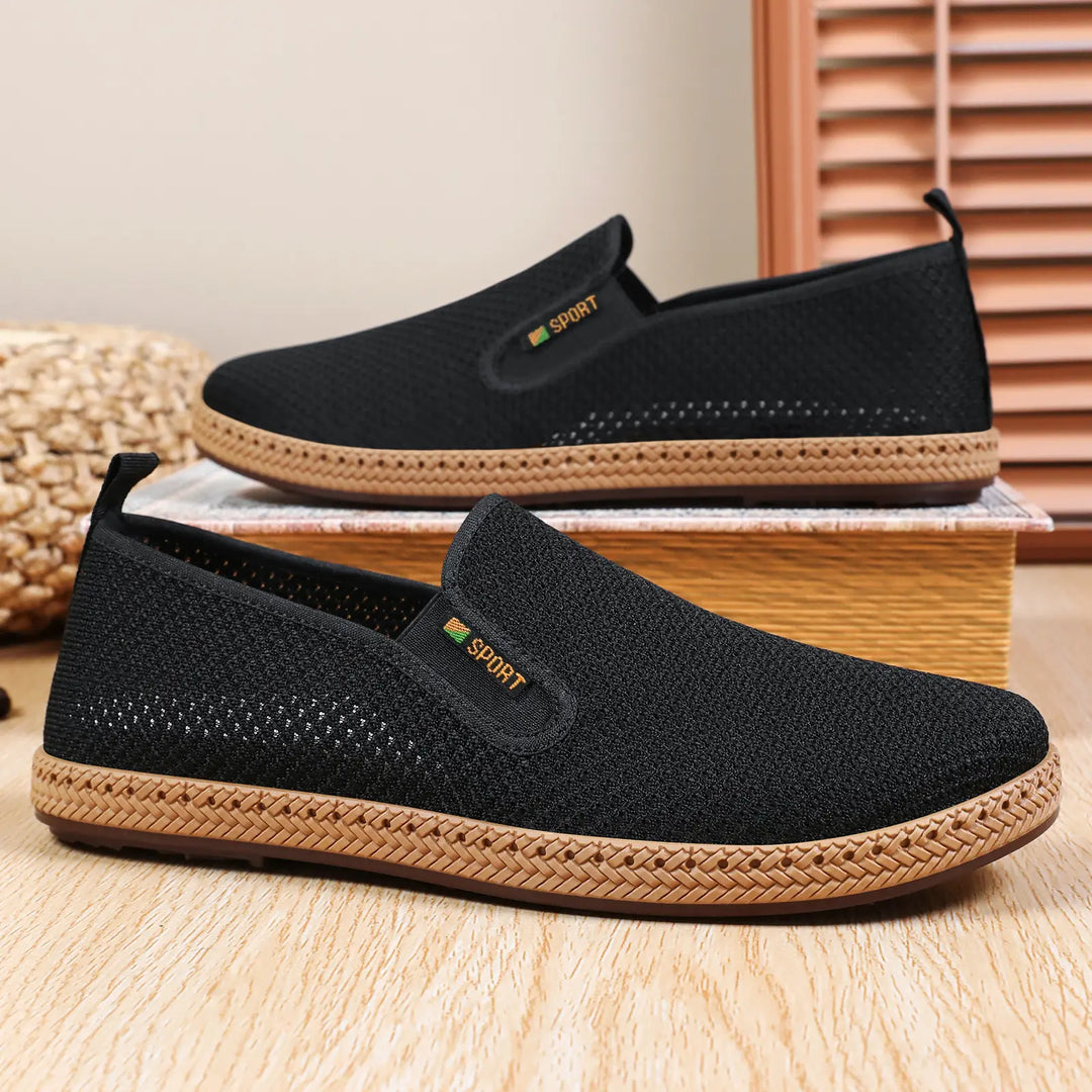 A pair of black breathable mesh slip-on loafers for men, shown from a three-quarter side view with woven jute-like trim and a subtle &