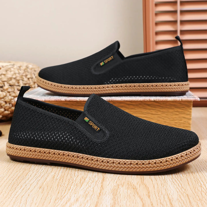 A pair of black breathable mesh slip-on loafers for men, shown from a three-quarter side view with woven jute-like trim and a subtle &