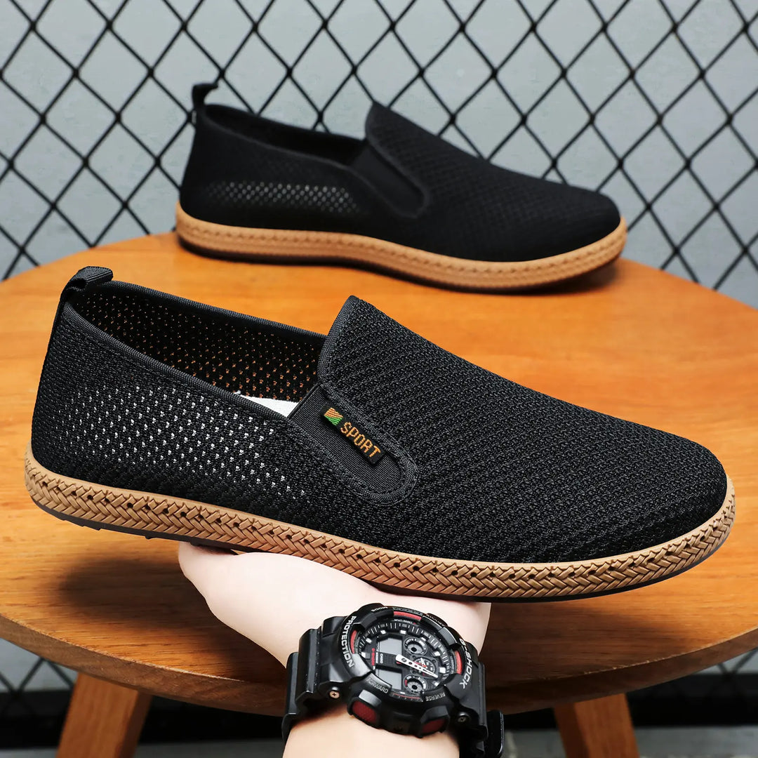 Black breathable mesh slip-on loafers with woven jute-like sole trim and a small "SPORT" logo accent on the side, displayed on a wooden surface.