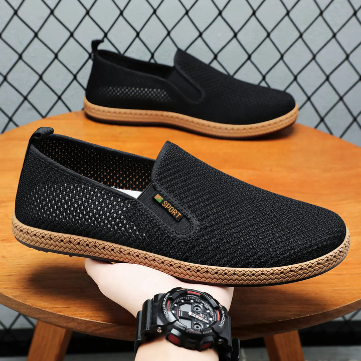 Black breathable mesh slip-on loafers with woven jute-like sole trim and a small "SPORT" logo accent on the side, displayed on a wooden surface.