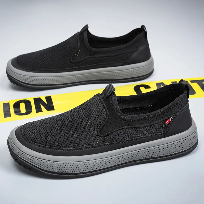 Pair of black breathable mesh slip-on sneakers with grey soles, viewed from a three-quarter angle in a studio setting with yellow cautionary tape behind them.