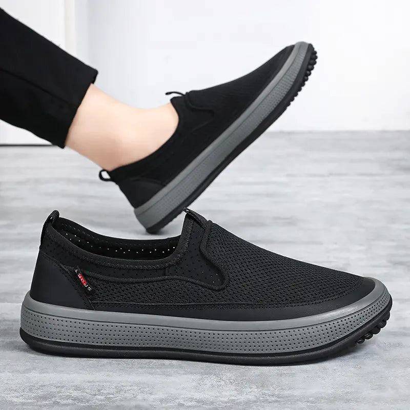 Black men's breathable mesh slip-on sneakers with a comfortable round toe, elastic gore panels, and durable grey rubber soles, shown from a three-quarter view with a secondary angle featuring a person's foot.