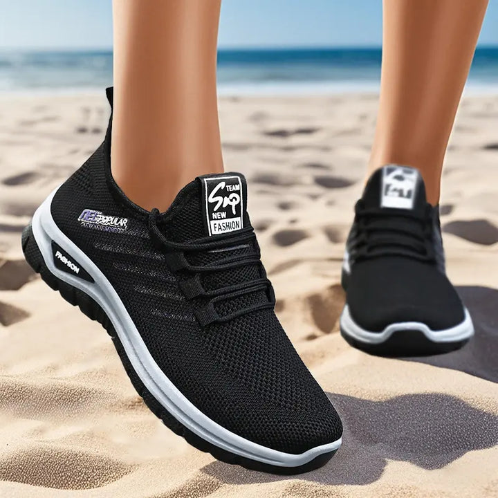 Pair of black breathable mesh sneakers worn on a sandy beach, showing the three-quarter front view of the left shoe with its white midsole and textured outsole.