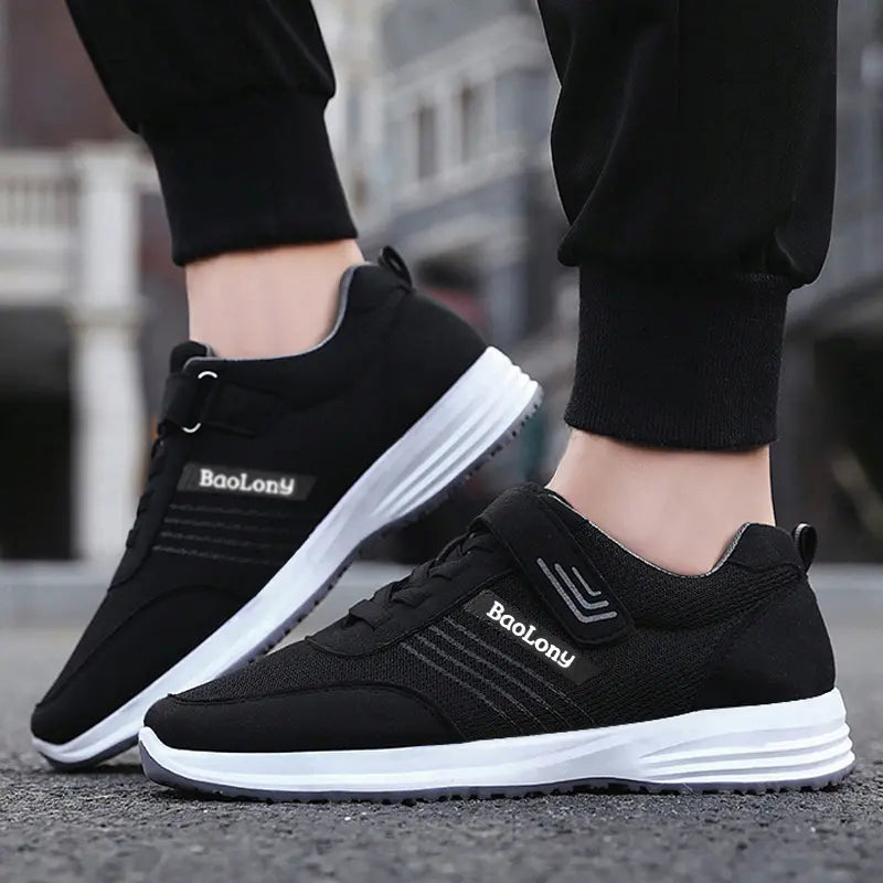 Black breathable mesh casual sneakers worn by feet, featuring a hook-and-loop strap, white midsole, grey outsole, and reflective side stripes.