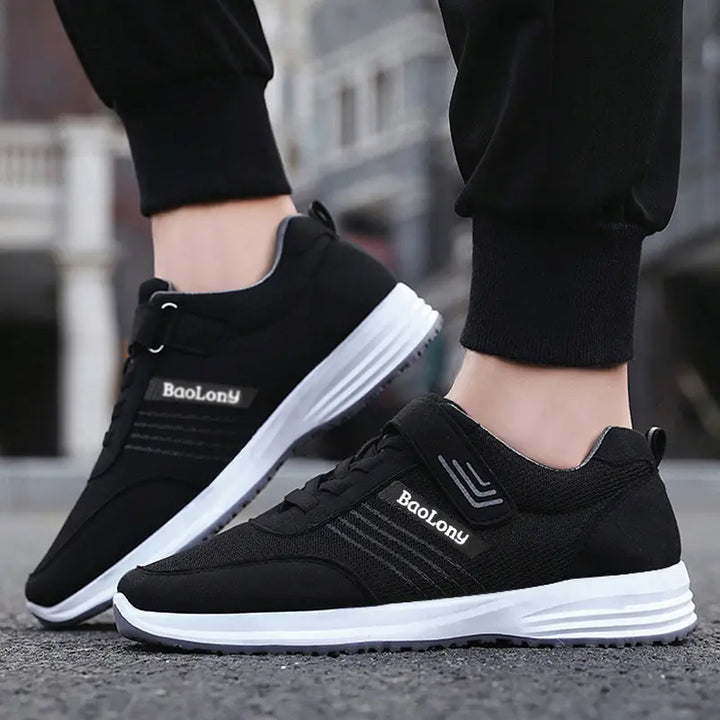 Black breathable mesh casual sneakers worn by feet, featuring a hook-and-loop strap, white midsole, grey outsole, and reflective side stripes.