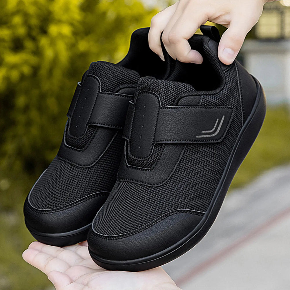 Black breathable mesh walking shoes with adjustable hook-and-loop straps, held by hands against a blurred green foliage background.