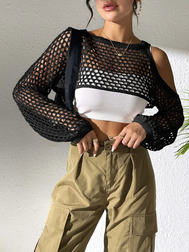 A model wears a black open-knit crochet crop top over a white ribbed bralette, styled with khaki cargo pants and gold jewelry against a textured wall background.