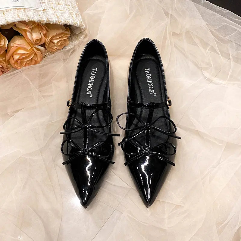 Pair of black patent leather pointed-toe ballet flats with decorative bows and adjustable ankle straps, presented on a draped fabric background.
