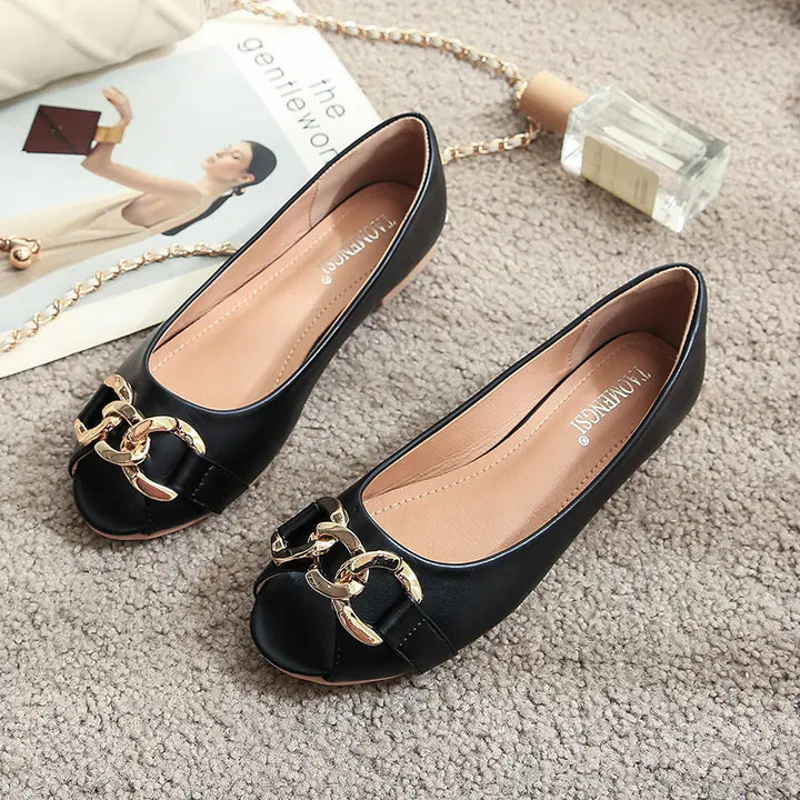 A pair of black faux leather peep-toe flats with gold chain embellishment, styled flat on a textured rug alongside a magazine and perfume bottle.
