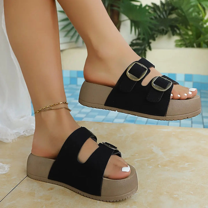 Black platform slide sandals with dual antiqued gold buckles and a beige sculpted sole, shown on feet in a summer lifestyle setting with a pool background.