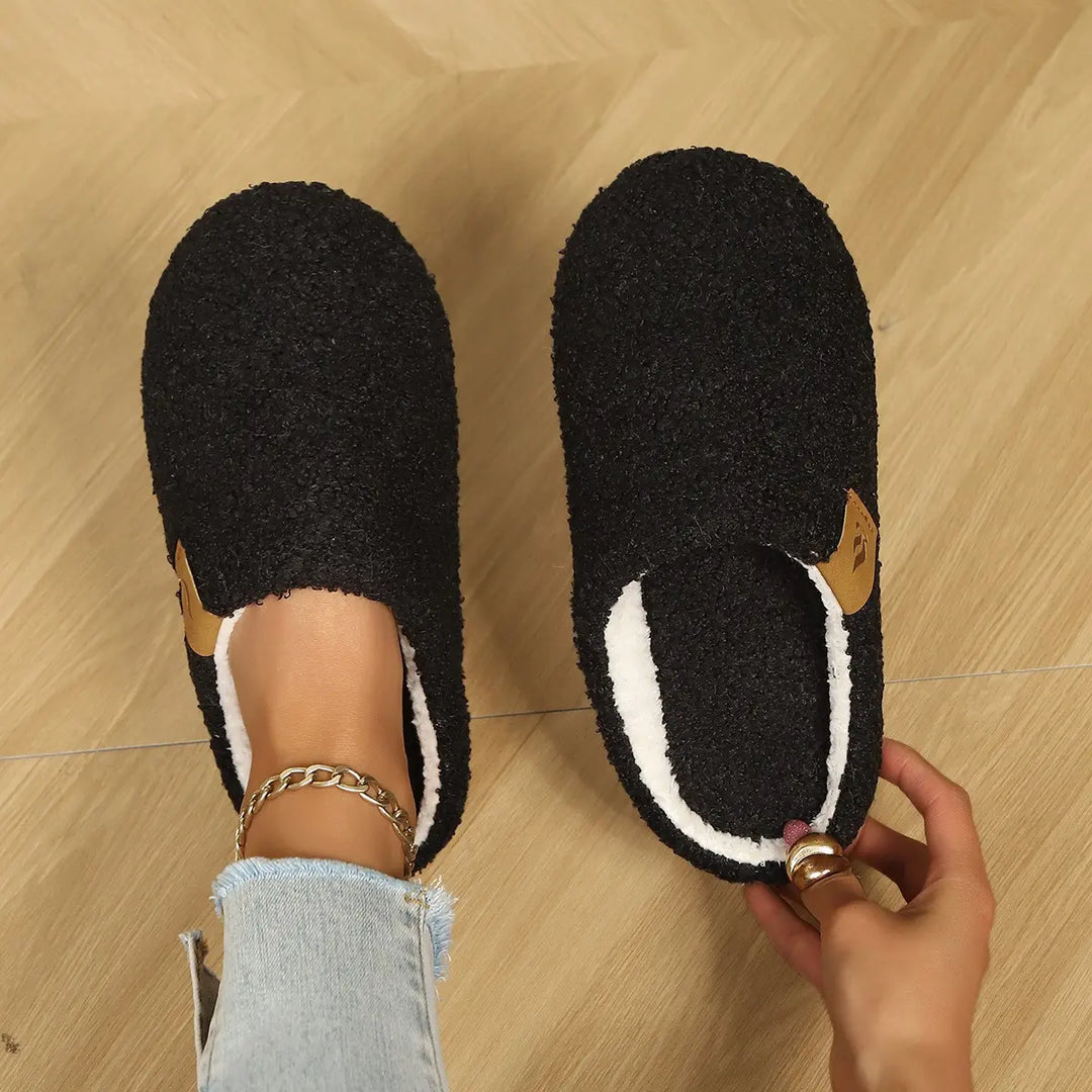 A pair of black plush clog slippers with white faux fur lining, worn by a person on a wooden floor, showing the faux leather accent tag. The wearer has on light-wash jeans and a gold chain anklet.