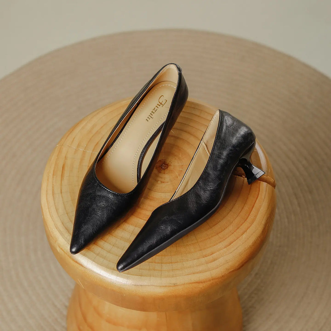 A pair of black faux leather pointed-toe pumps with a flared kitten heel, presented on a wooden stool in a studio setting.
