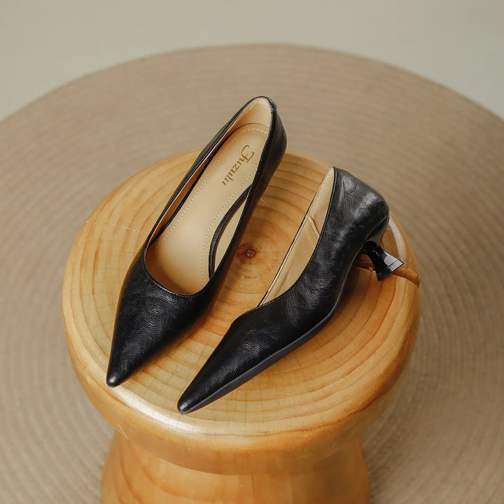 A pair of black faux leather pointed-toe pumps with a flared kitten heel, presented on a wooden stool in a studio setting.