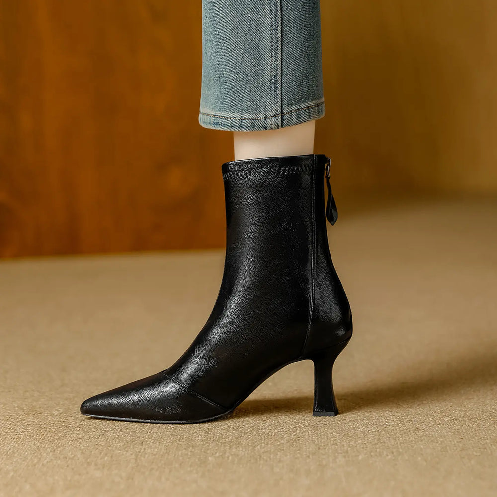 Black faux leather pointed toe ankle boot with a stiletto heel and back zipper, styled with denim jeans on a neutral studio floor.