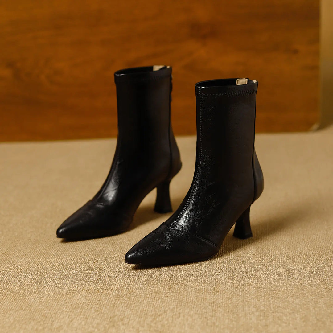 Black faux leather pointed toe stiletto ankle boots, presented from a front angled view on a textured floor with a wooden background.