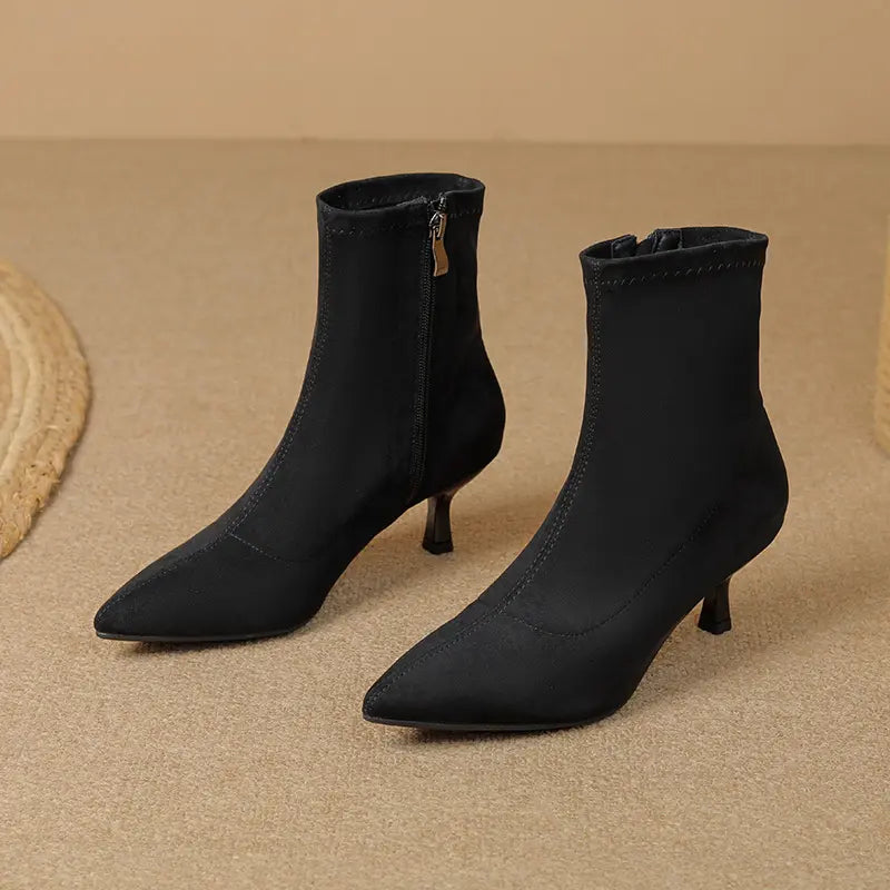 Pair of black suede-like stiletto ankle boots with pointed toes and inner side zippers, shown from a three-quarter angle on a textured studio background.