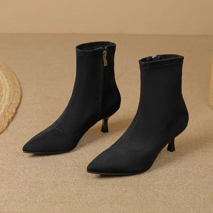 Pair of black suede-like stiletto ankle boots with pointed toes and inner side zippers, shown from a three-quarter angle on a textured studio background.