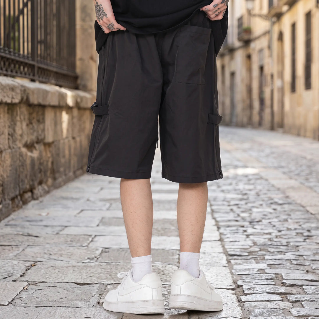 Matte black relaxed-fit technical shorts with diagonal zip pockets, elasticated waistband, and rear snap button pockets, shown on a model from the front on a white studio background.