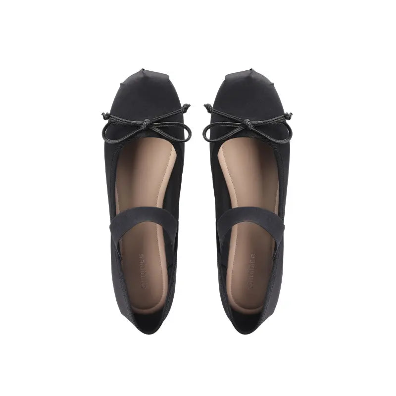 A pair of black satin ballet flats with a delicate bow on the vamp and a visible elastic instep strap, shown from a top-down view on a white background.