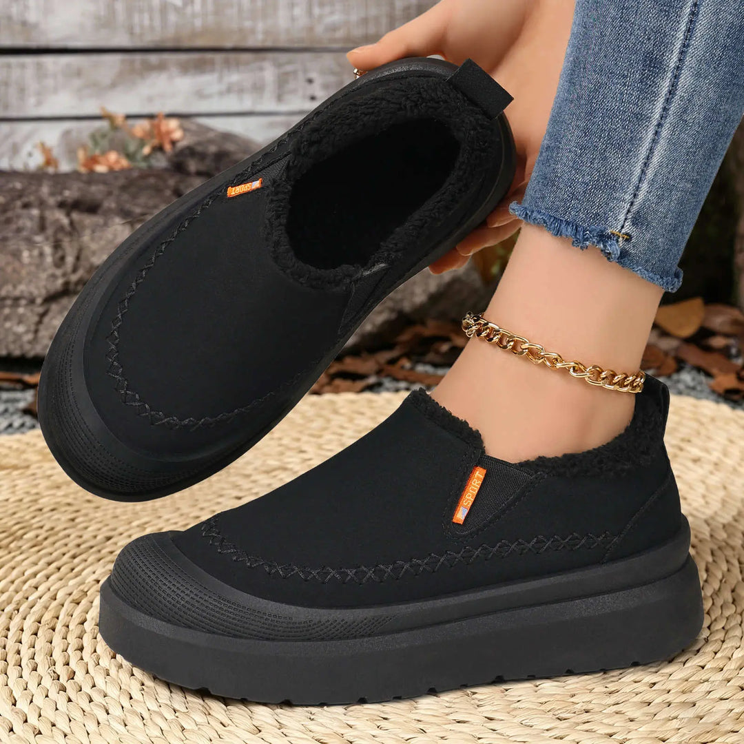 Black slip-on comfort boots with plush shearling lining and decorative stitching, shown on a person&
