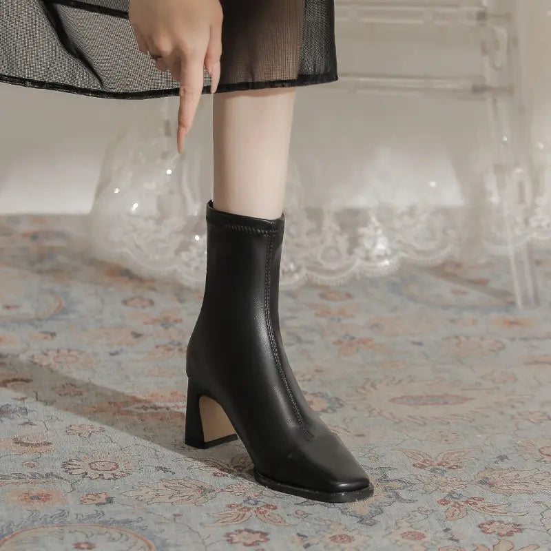 Black faux leather ankle boot with a chic square toe and stable block heel, worn with a skirt for a stylish seasonal look.