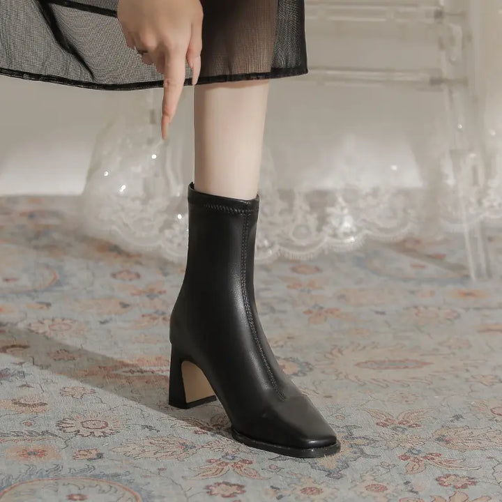 Black faux leather ankle boot with a chic square toe and stable block heel, worn with a skirt for a stylish seasonal look.