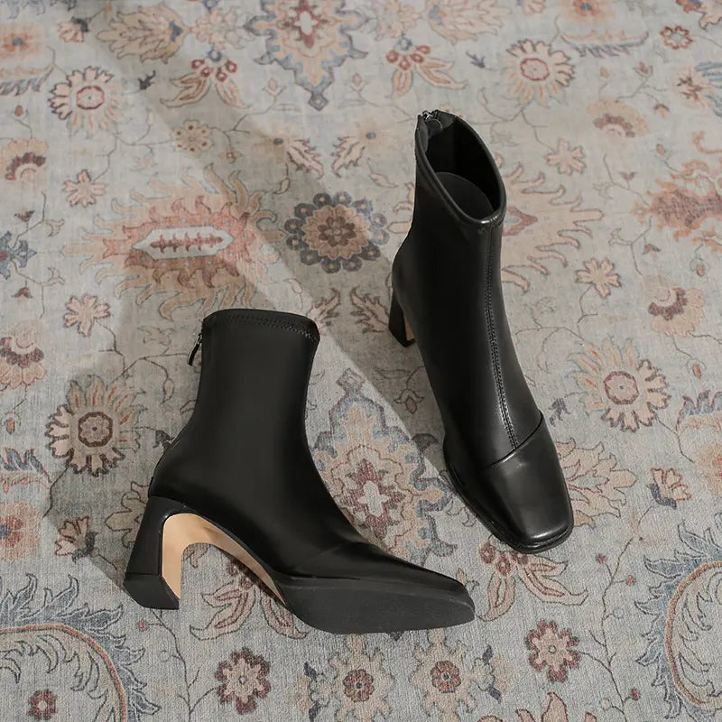 Chic black faux leather ankle boots with a square toe and block heel, presented on a patterned rug. These stylish boots feature an interior side zipper and a mid-calf shaft.