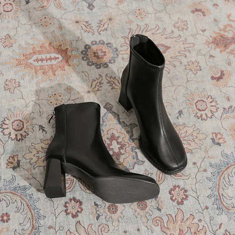 Black faux leather ankle boots featuring a contemporary square toe, chunky block heel, and full-length back zipper, shown from a three-quarter view on a patterned rug.