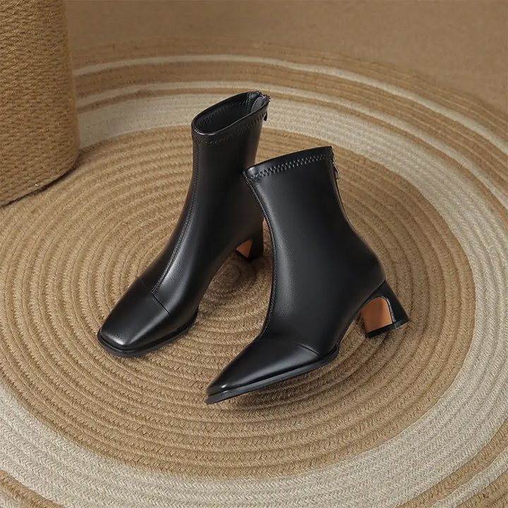 Chic black faux leather ankle boots featuring a sharp square toe, interior side zipper, and a distinctive geometric block heel, presented on a textured woven rug.