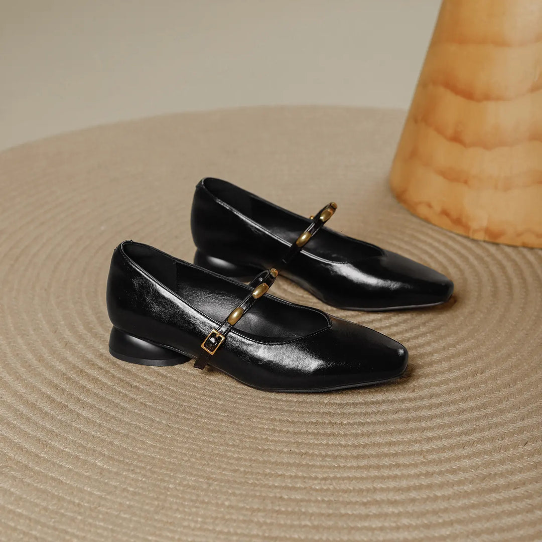 Black high-shine microfiber square toe Mary Jane shoes with a low block heel and gold-tone buckle strap, shown from a three-quarter angle on a textured rug.
