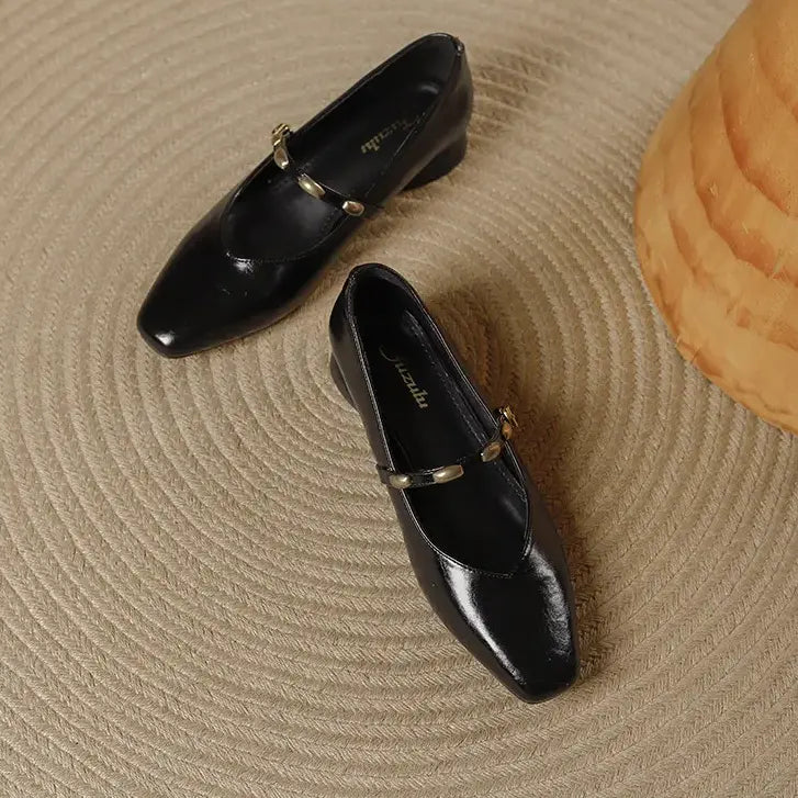 Pair of black high-shine microfiber square-toe Mary Jane shoes with gold-tone buckle straps, shown from a top-down angle on a textured rug.
