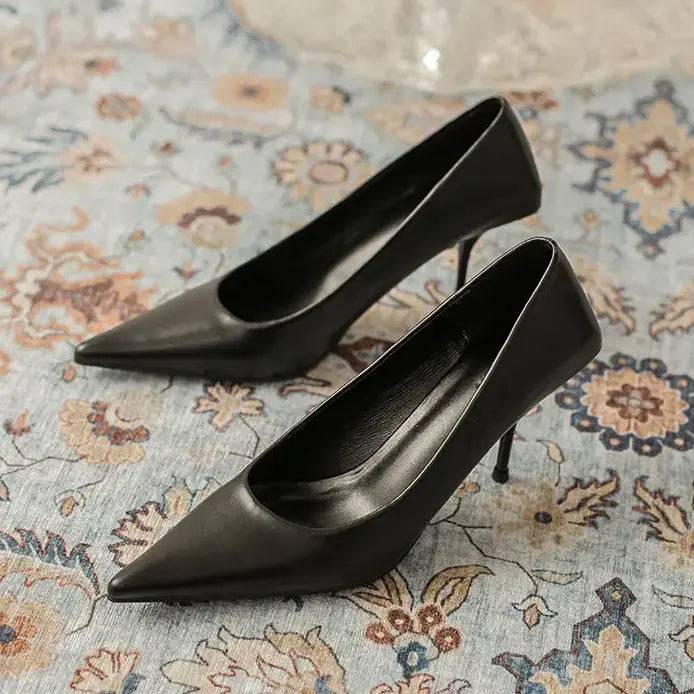 Black high-gloss faux leather stiletto heels with sharp pointed toes, resting on a decorative patterned rug.