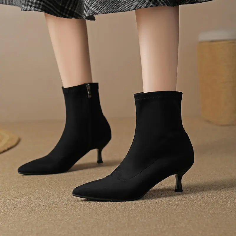 Black suede-like pointed-toe stiletto ankle boots with a stretch sock shaft and inner side zipper, shown worn with a skirt.