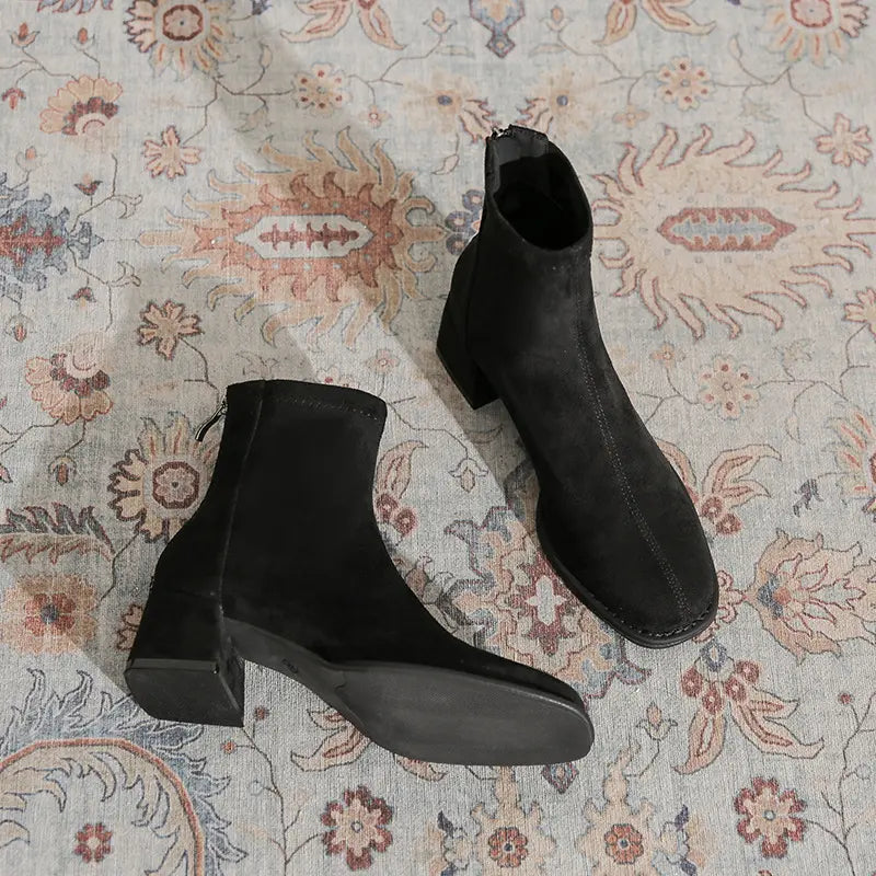 Black suede-like ankle boots with a square toe, chunky block heel, and back zipper, shown from a three-quarter angle on a patterned rug.