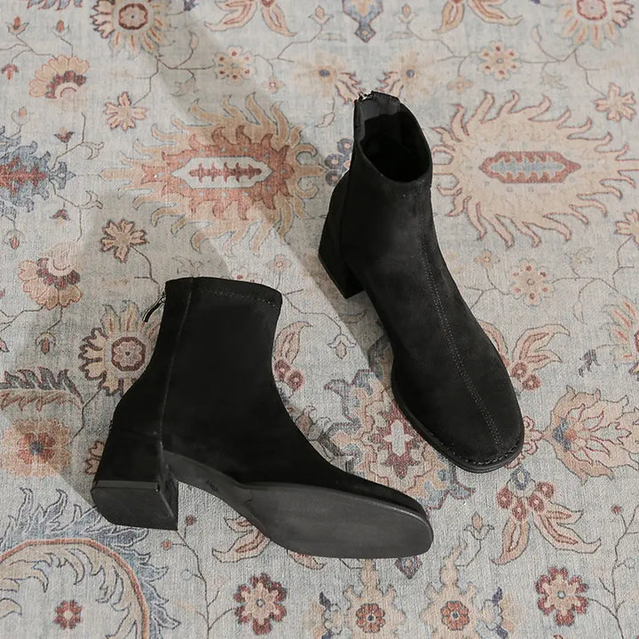 Black suede-like ankle boots with a square toe, chunky block heel, and back zipper, shown from a three-quarter angle on a patterned rug.