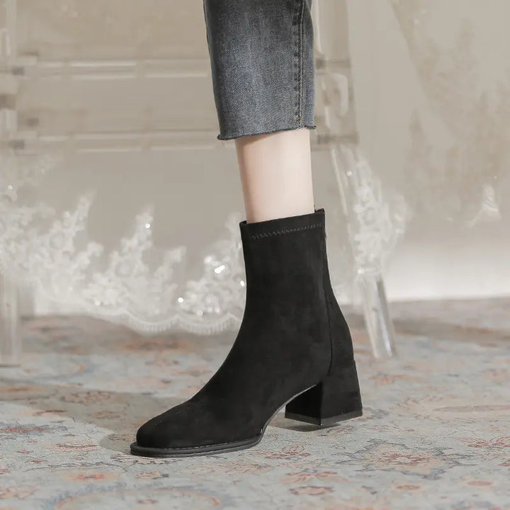 A pair of black suede-like ankle boots with a square toe and block heel, worn with cropped denim jeans on a patterned rug, showcasing a chic winter footwear style.