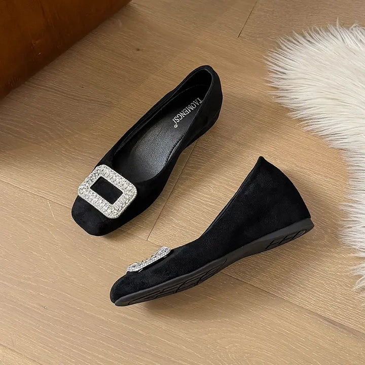 Black suede slip-on wedge flats with a rounded-square toe and a dazzling crystal-embellished square buckle on the vamp, displayed on a wooden floor.