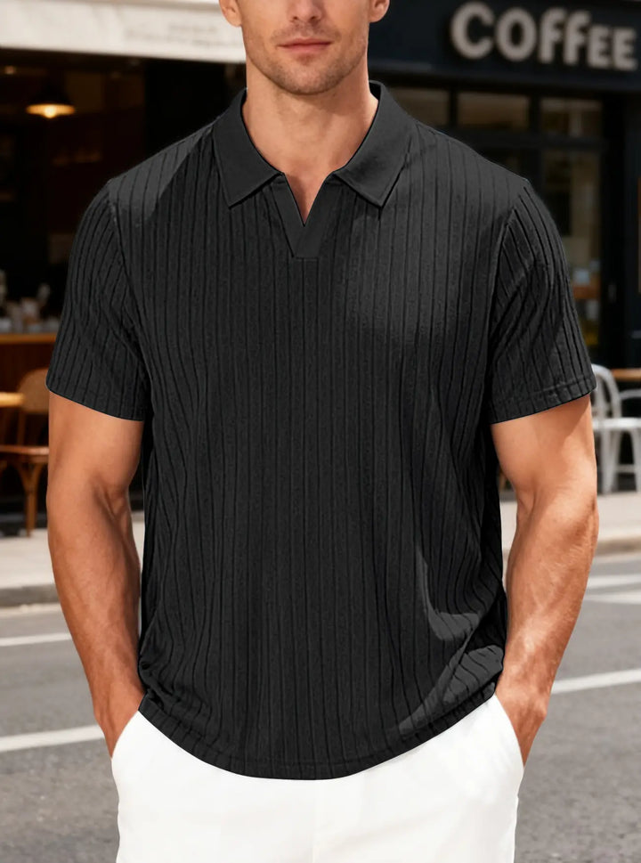 Man wearing a black textured knit short sleeve V-neck polo shirt with fine vertical ribbing, styled with white pants in an outdoor lifestyle setting.