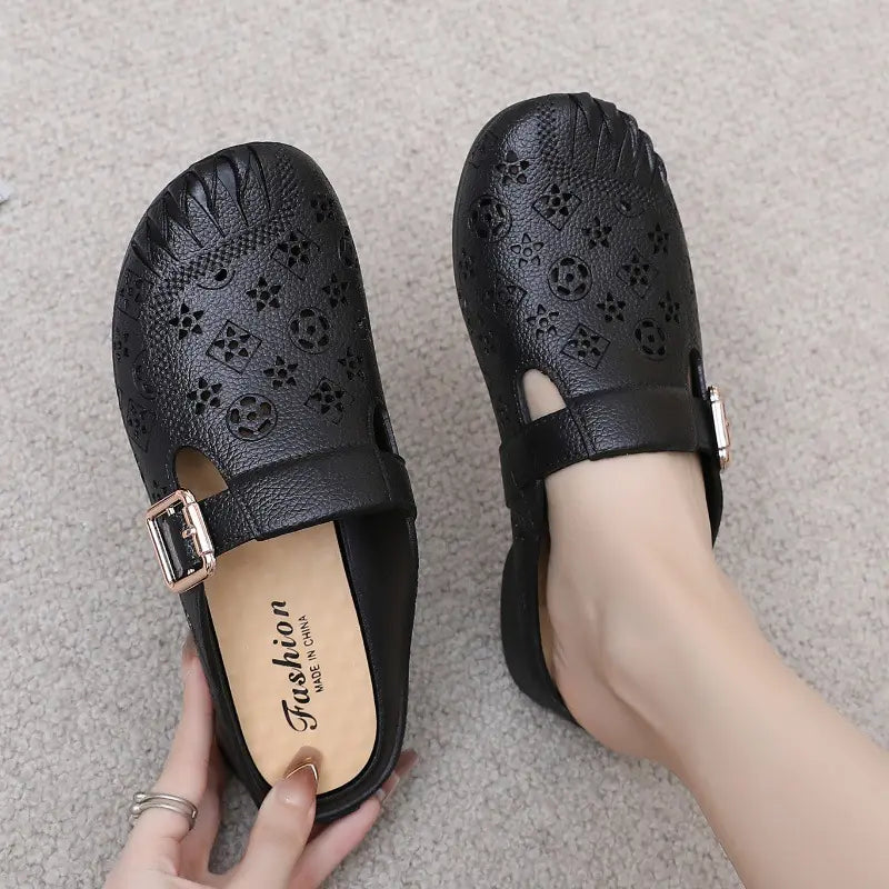 Black textured synthetic mules with an embossed geometric pattern and an adjustable gold-tone buckle strap, shown on a foot with the other shoe held, on a carpeted background.