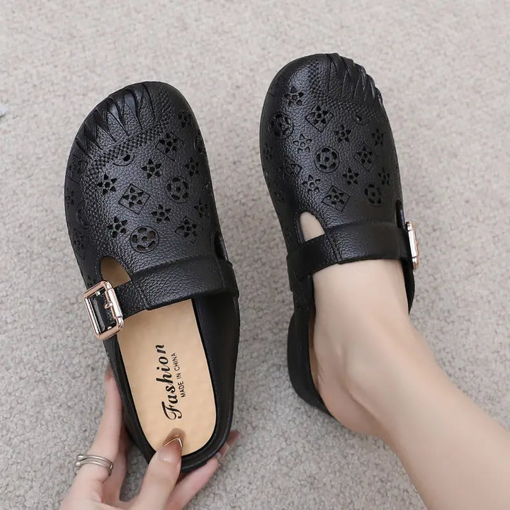 Black textured synthetic mules with an embossed geometric pattern and an adjustable gold-tone buckle strap, shown on a foot with the other shoe held, on a carpeted background.